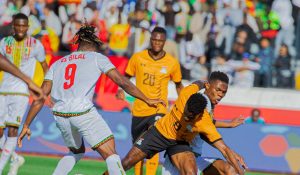 Action shot of a Mali player in a white jersey (number 9, El Bilal) and two Zambia players in orange jerseys competing for the ball.
