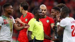 A referee in a yellow Puma kit surrounded by several football players in red jerseys and white jerseys, all appearing to engage in an intense discussion on the pitch.