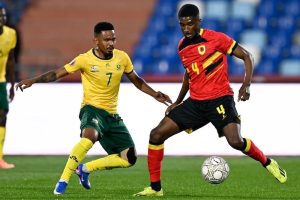 Action shot of an Angolan soccer player in a red jersey (number 4) shielding the ball from a South African player in a yellow jersey (number 7).