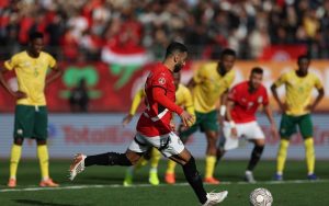 Egyptian football player Mohamed Salah in a red jersey taking a penalty kick while South African players in yellow jerseys watch from the background.