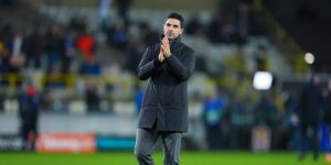 A medium shot of Arsenal manager Mikel Arteta standing on a professional football pitch at night. He is wearing a dark, quilted winter coat over grey trousers and is holding his hands together in a clapping or thoughtful gesture. The background shows the blurred stands of a stadium with spectators and bright stadium lights.