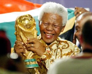 A joyful Nelson Mandela wearing a patterned gold shirt and holding the gold FIFA World Cup trophy aloft, with the South African flag visible in the background.
