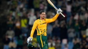 South African cricketer Quinton de Kock raising his bat and helmet in celebration, wearing a yellow and green Proteas limited-overs jersey in a crowded stadium.